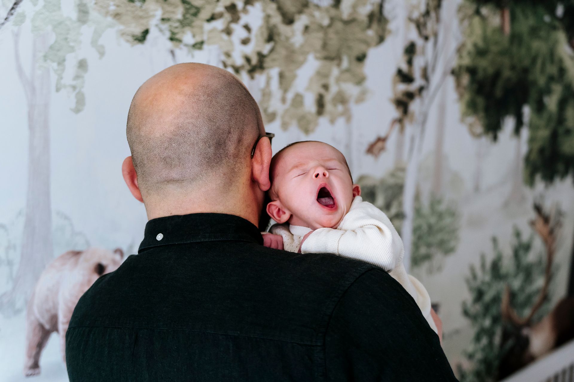 Een man draagt ​​een gapende baby over zijn schouder in een kinderkamer met behang met een bosmotief.
