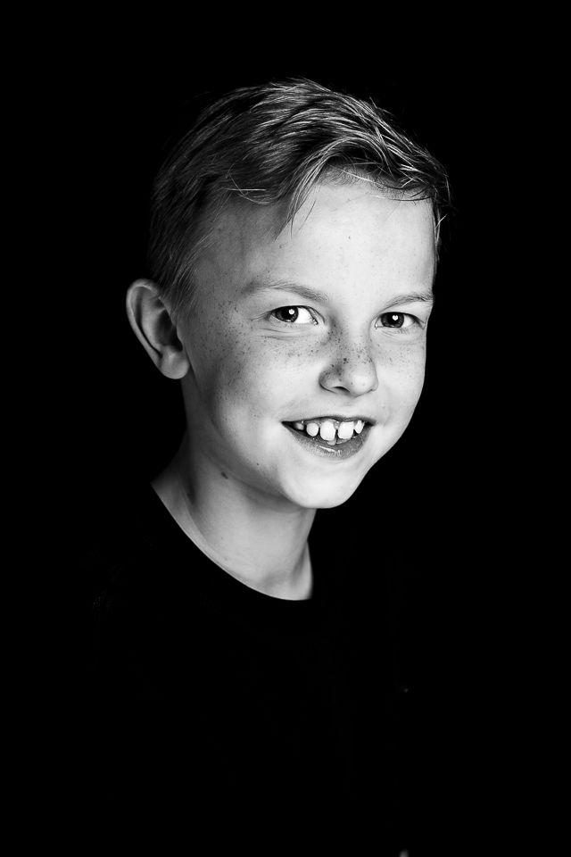 Jongen lacht en kijkt naar de camera. Zwart-wit, tegen een donkere achtergrond.
