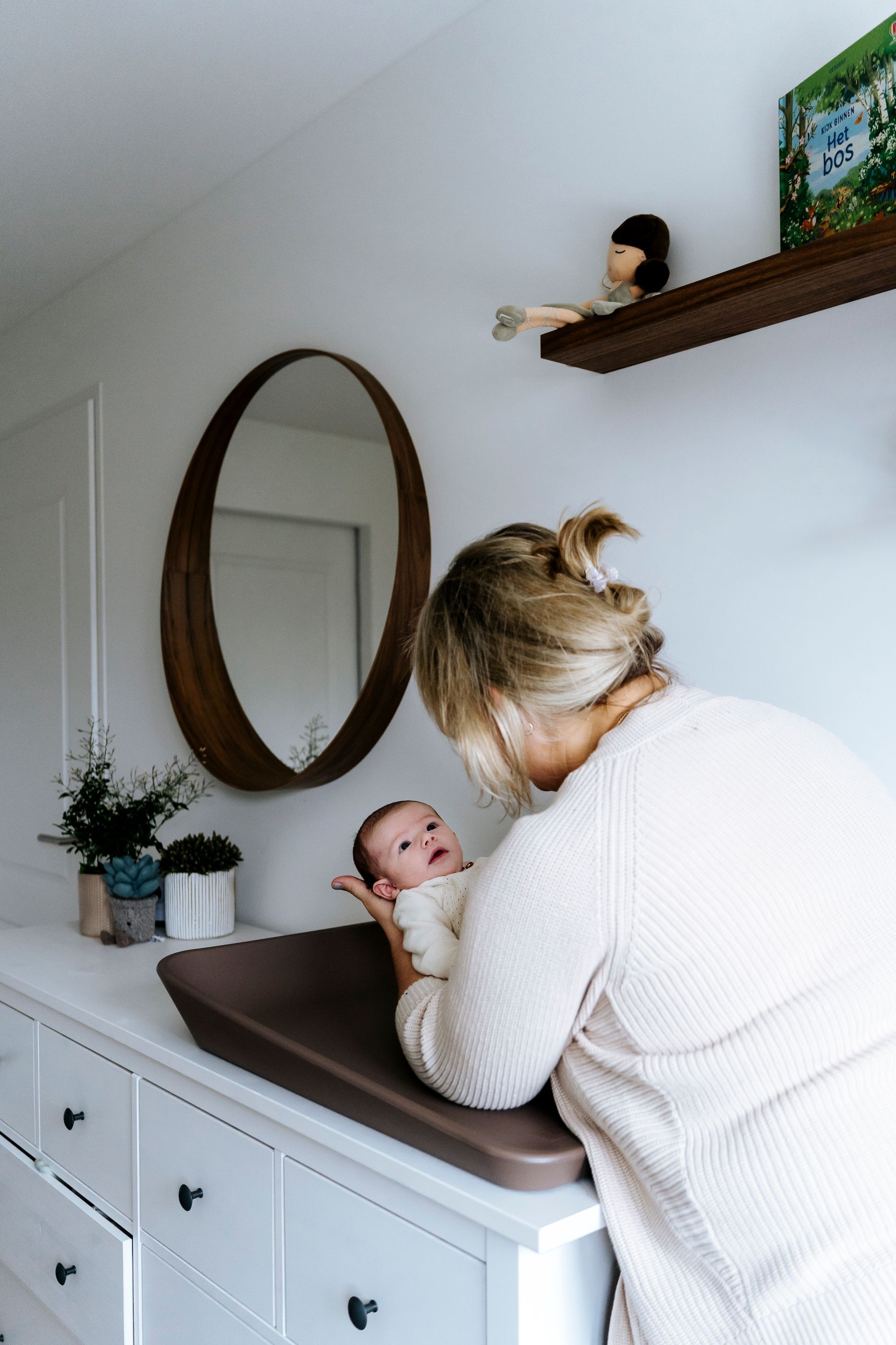 Vrouw met baby op een commode, kijkt naar beneden. Kamer met spiegel en planken.