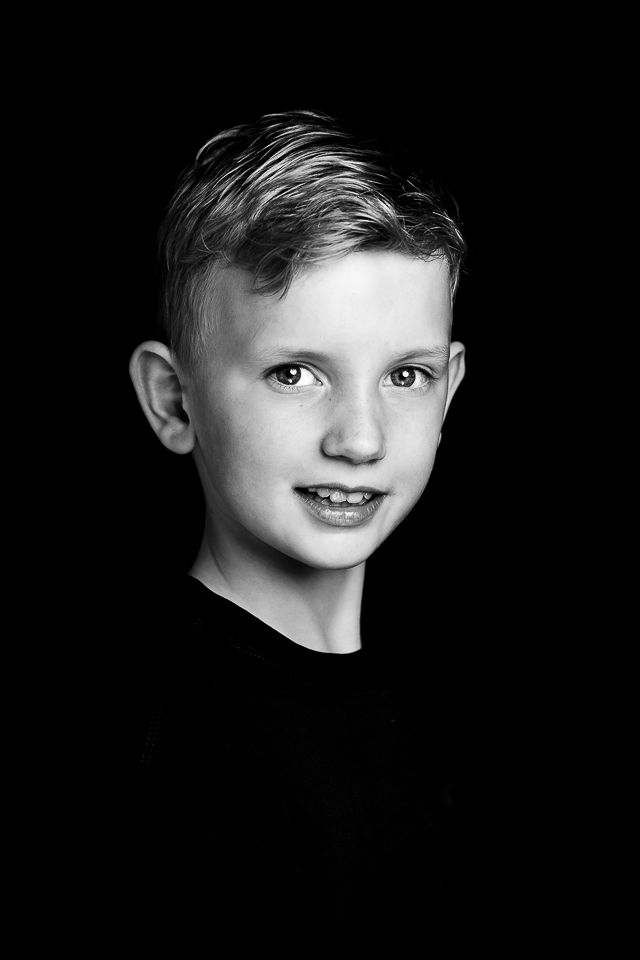 Jongen met blond haar, lachend. Zwart-wit portret tegen een zwarte achtergrond.