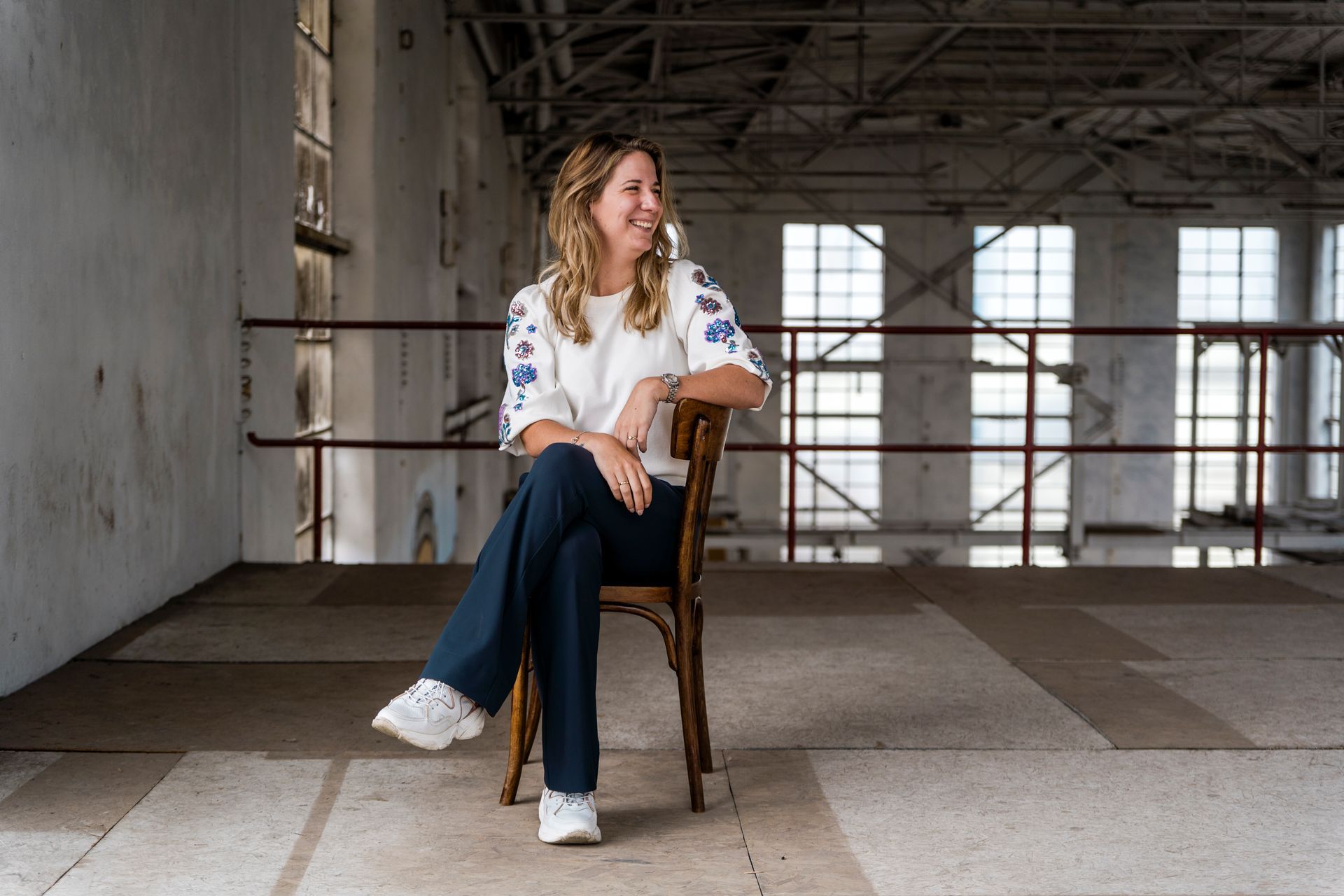 Een vrouw in een witte, geborduurde top en een blauwe broek zit op een stoel in een industriegebouw, glimlachend en opzij kijkend.