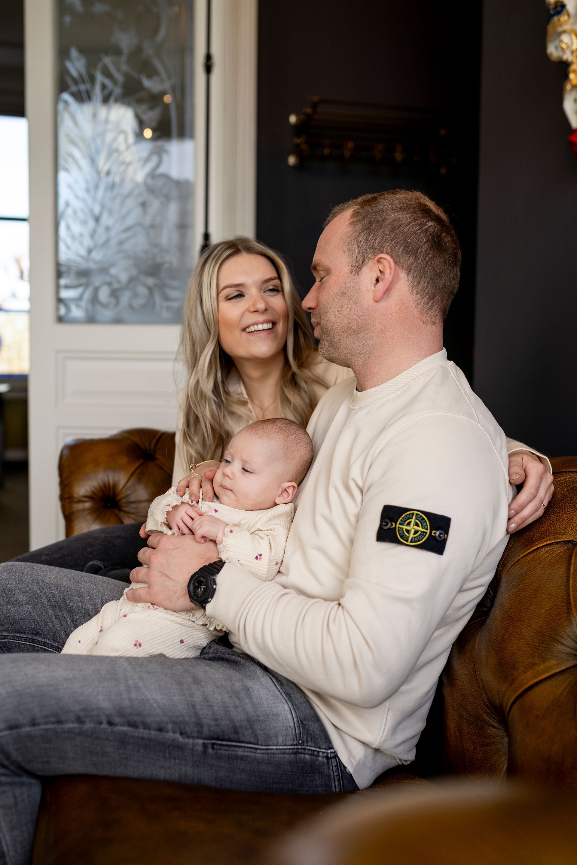 Een gezin van drie zit op een bruine leren bank; de vrouw glimlacht, de man houdt een baby vast en kijkt elkaar aan.