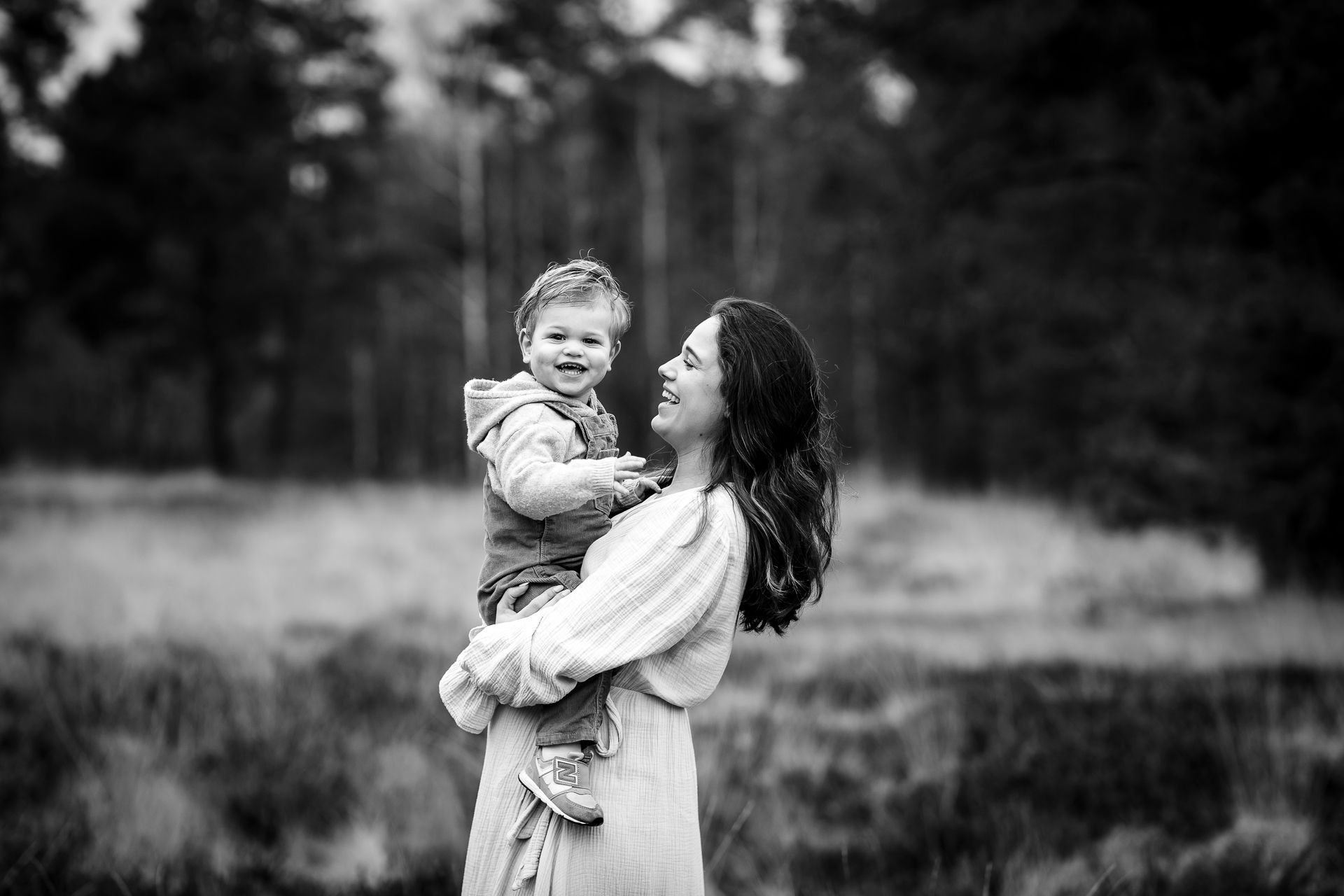 Een vrouw houdt een kind vast en lacht ernaar in een veld, met bomen op de achtergrond. Zwart-witfoto.