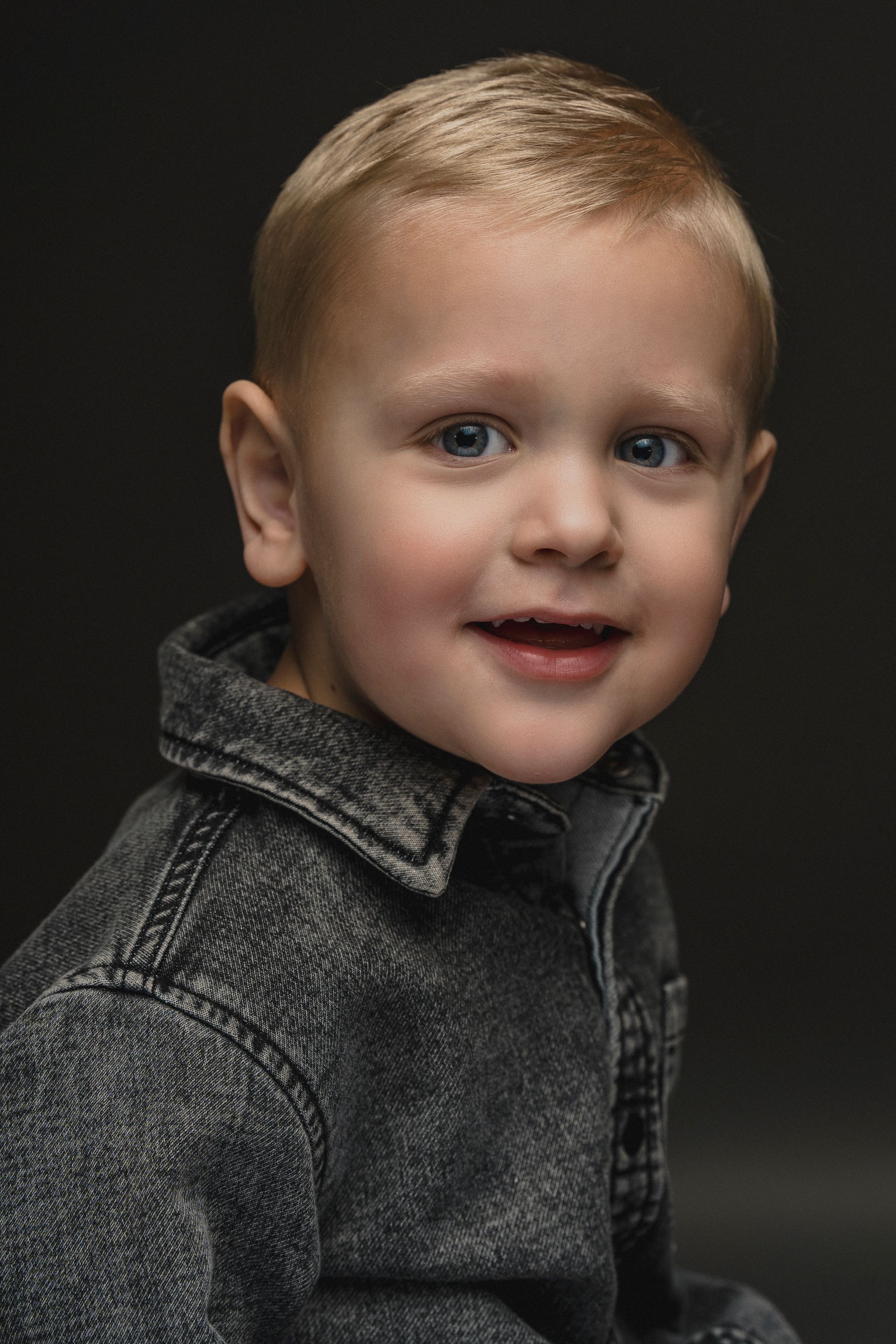 Een blond kind lacht, draagt een donker spijkerjack en staat voor een donkere achtergrond.