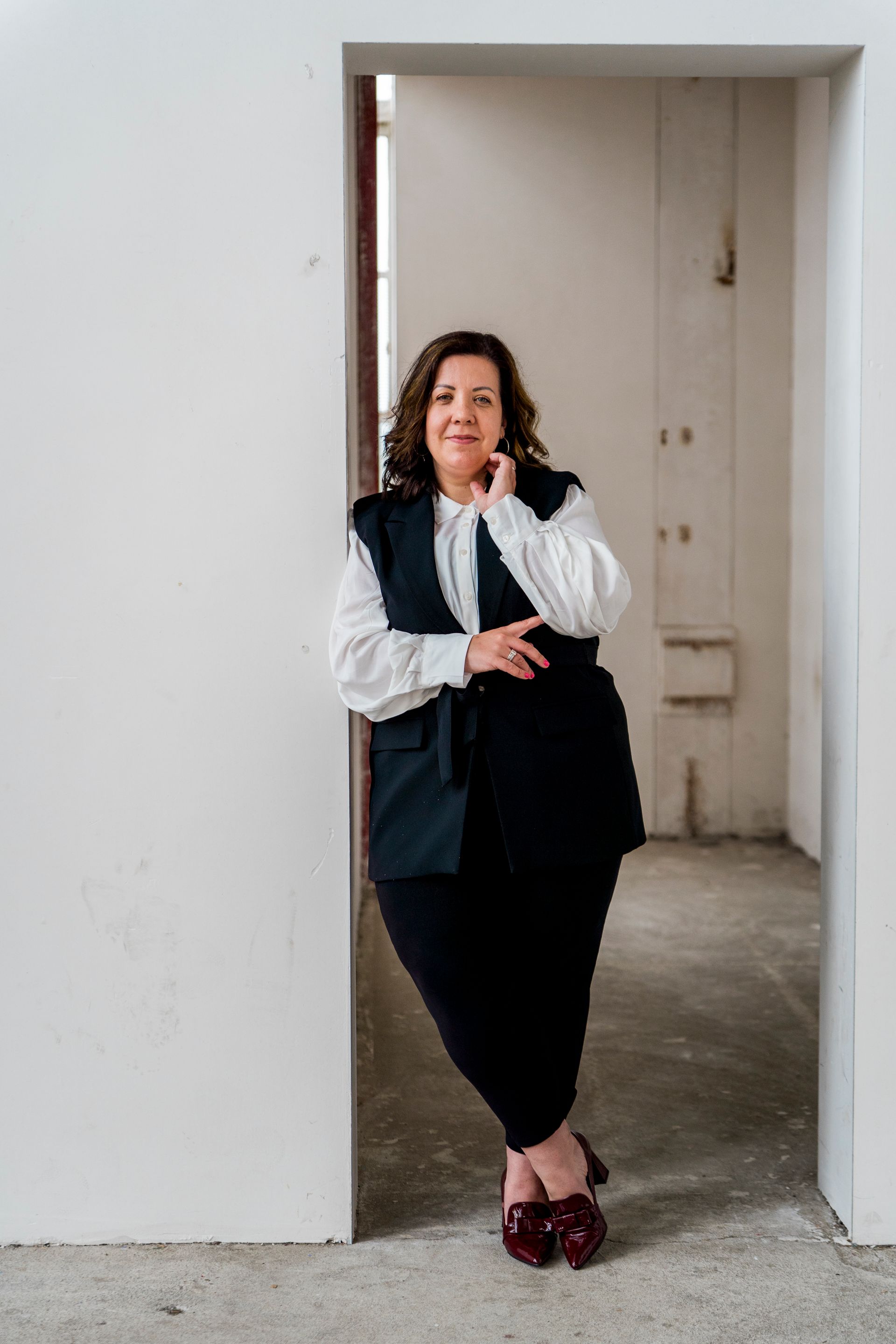 Een vrouw in een zwart vest en rok leunt tegen een witte muur in een deuropening.