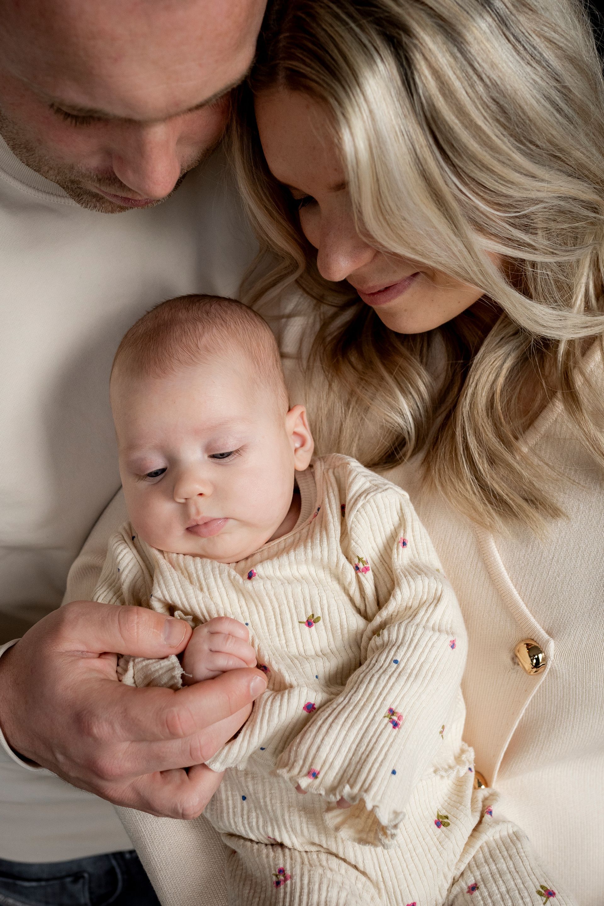 Ouders houden hun baby liefdevol vast, gekleed in crèmekleurige gebreide kleding. De baby kijkt naar beneden.
