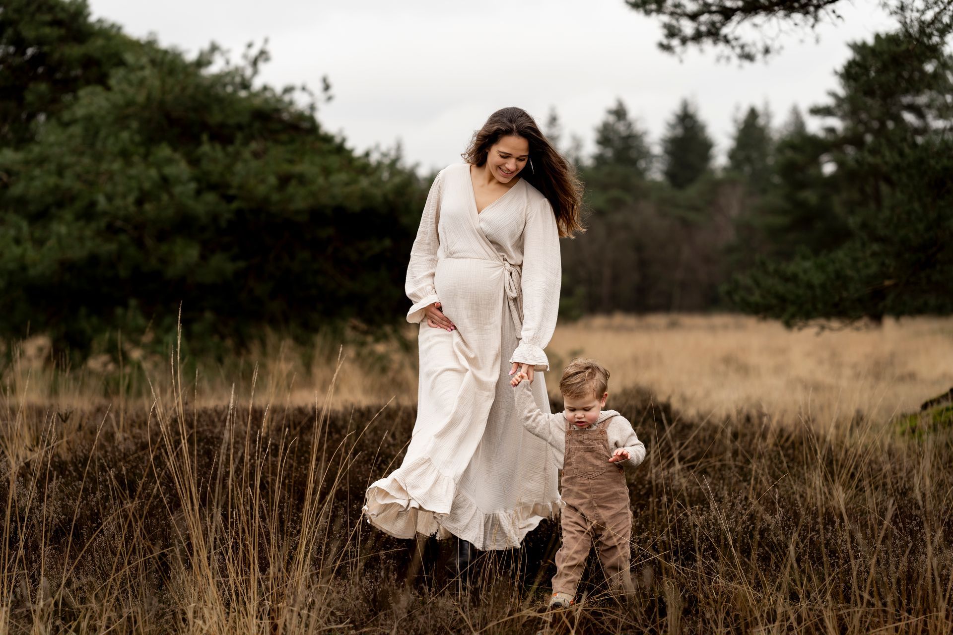 Een zwangere vrouw en haar peuter lopen door hoog gras in een veld, met bomen op de achtergrond.