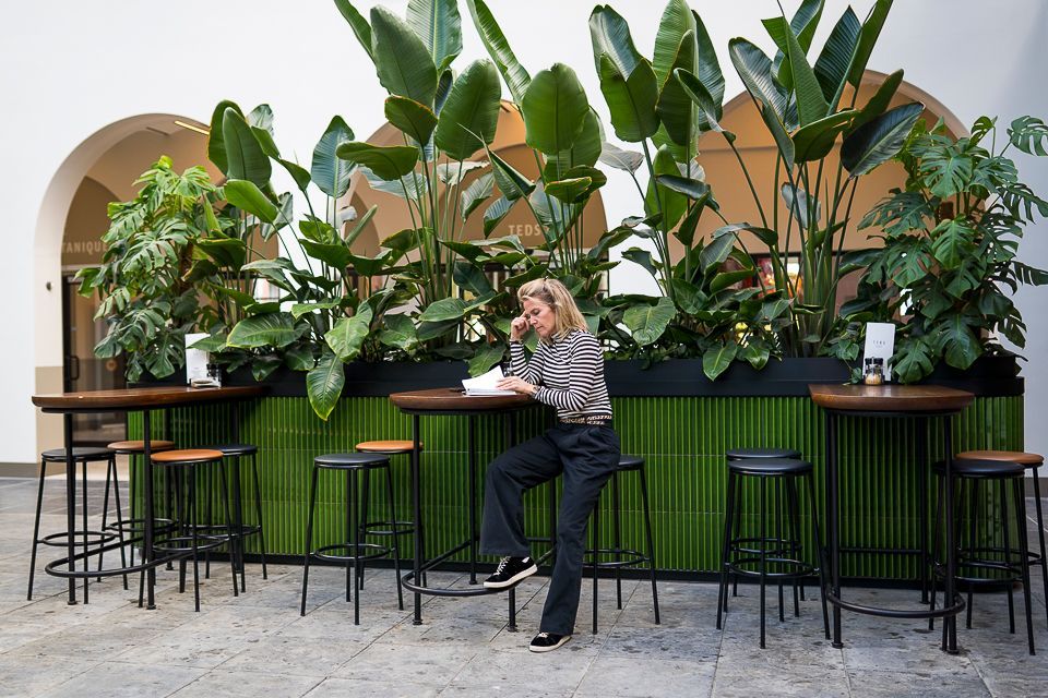 Een vrouw zit aan een tafel aantekeningen te maken, omringd door weelderige planten en barkrukken in een lichte, moderne ruimte.