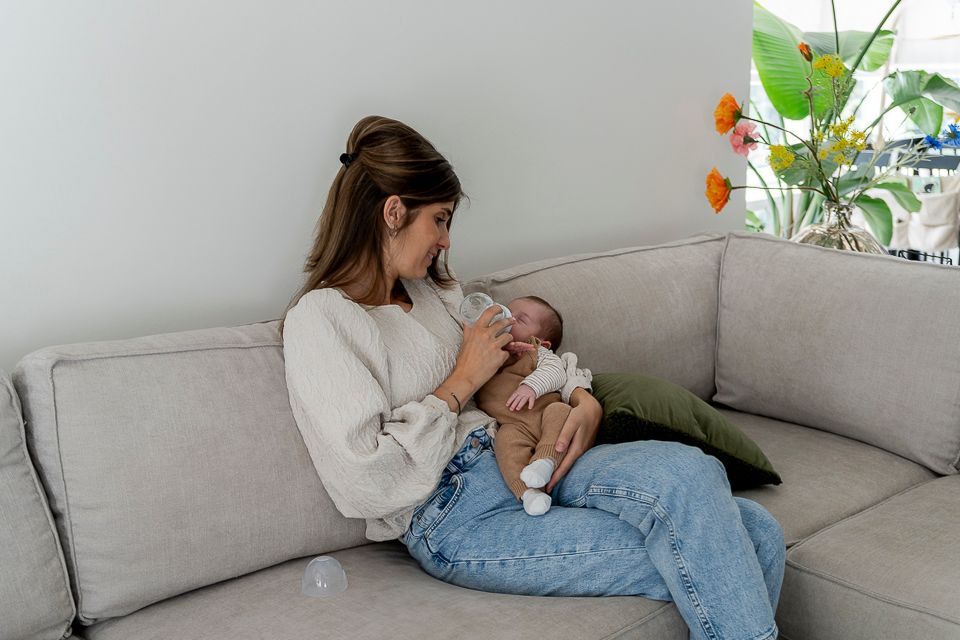 Een vrouw geeft een baby de fles op een grijze bank in een kamer vol bloemen.