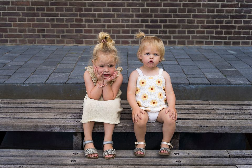 Twee jonge kinderen zitten somber op een houten bankje. De ene draagt ​​crèmekleurige kleding, de andere een outfit met bloemenprint. Op de achtergrond een bakstenen muur.