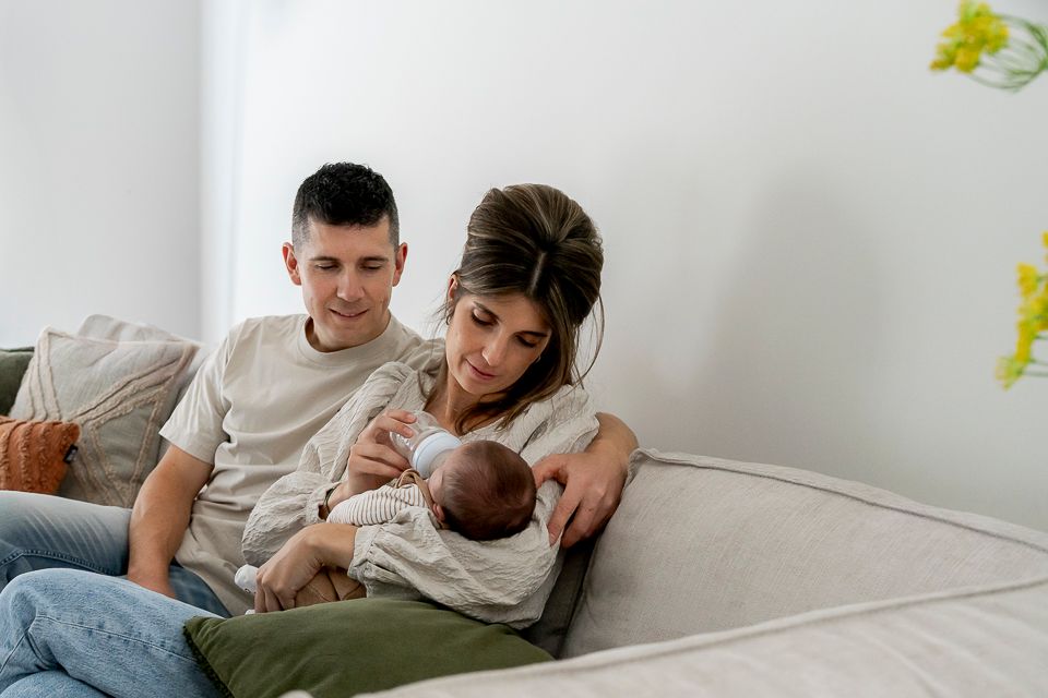 Ouders geven hun baby de fles op de bank. De vrouw houdt de baby vast, de man kijkt toe. Woonkamer, neutrale kleuren.