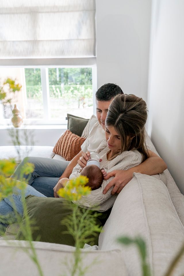 Ouders wiegen hun pasgeboren baby op een bank bij een raam; natuurlijk licht, neutraal kleurenpalet.