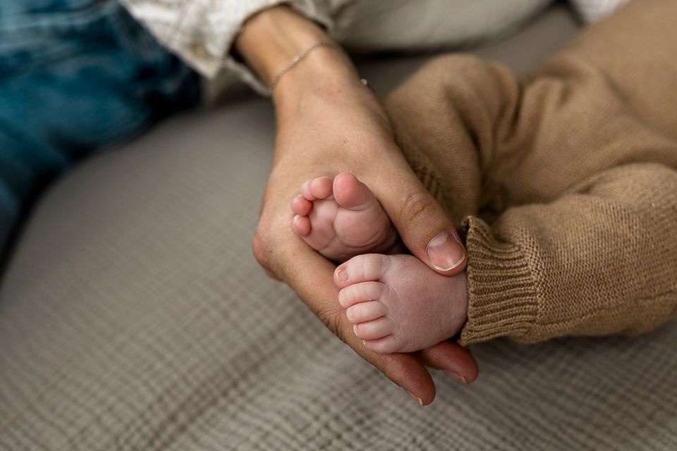 De hand van een ouder ondersteunt de voetjes van de baby, die een beige romper draagt en op een deken in een neutrale kleur ligt.