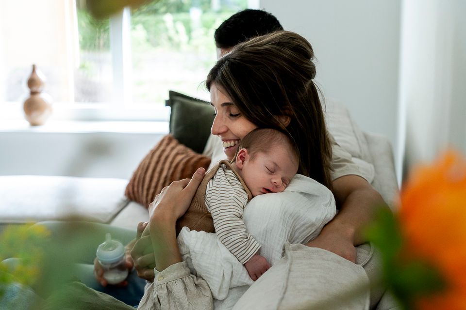 Een moeder wiegt een slapende baby in haar armen, terwijl de vader zijn arm om hen heen slaat op een bank; binnenscène, natuurlijk licht.