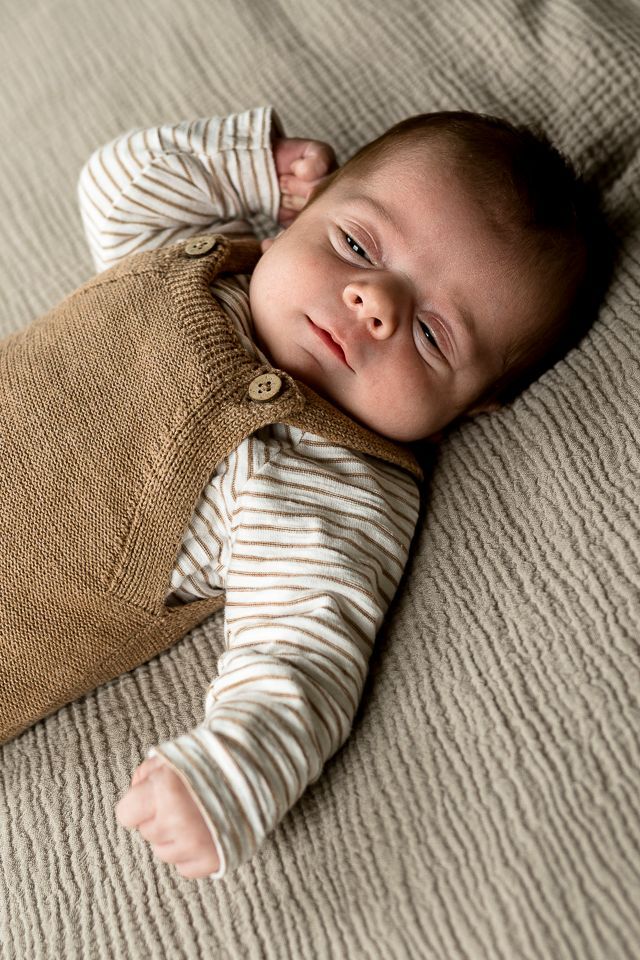 Baby in bruine gebreide tuinbroek en gestreept shirt, liggend op een grijze deken met structuur.