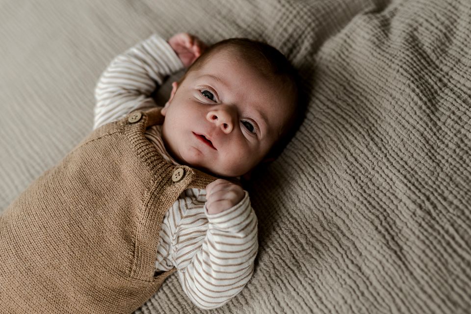 De baby ligt op een grijze deken met structuur, gekleed in een bruin gebreid pakje en een gestreept shirt, en kijkt omhoog.