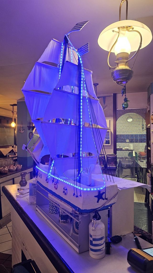 Esposizione al chiuso di un modellino di barca a vela illuminato di blu, con vele gonfiate dal vento e una lanterna accanto.