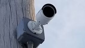 Security camera mounted on a wooden pole, white camera, gray box, sky in background.