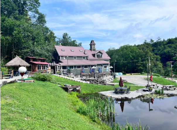 Green building with mini golf course, pond, and outdoor seating on a sunny day.