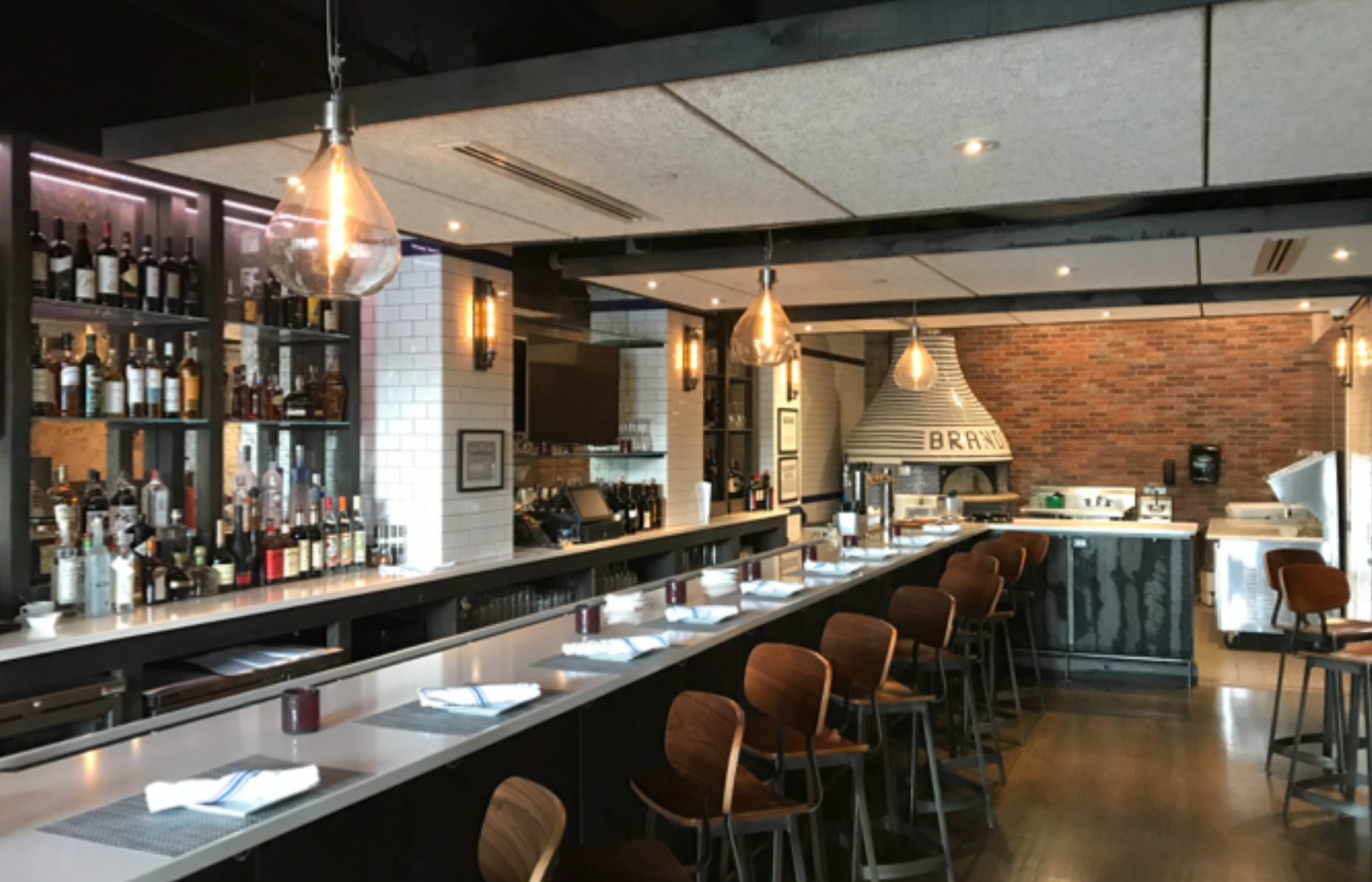 Restaurant interior with bar, brick oven, and seating. Dark wood bar and stools, exposed brick, and pendant lights.