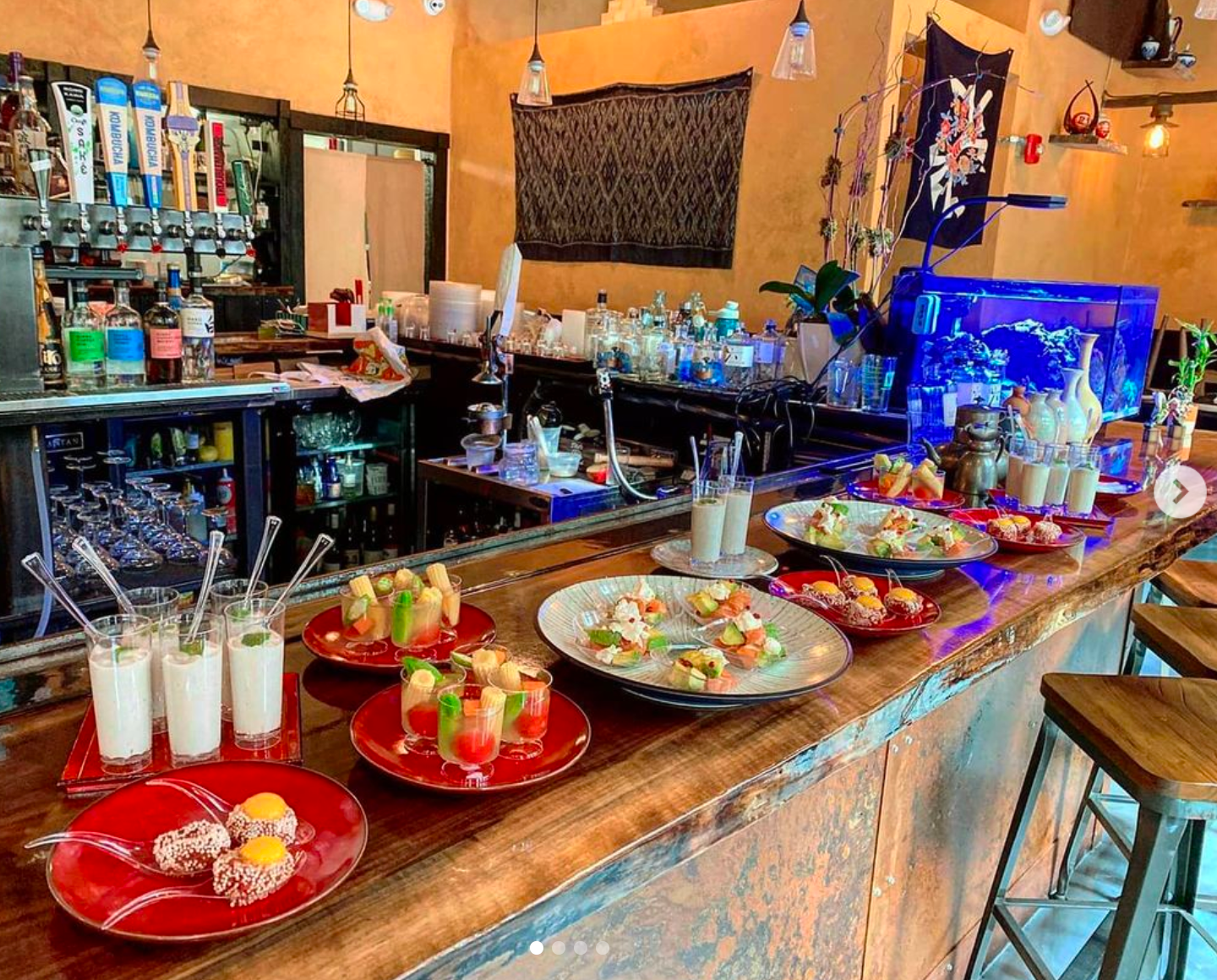 A bar counter displays prepared food and drinks. Wooden stools line the front. A bar and aquarium are in the background.