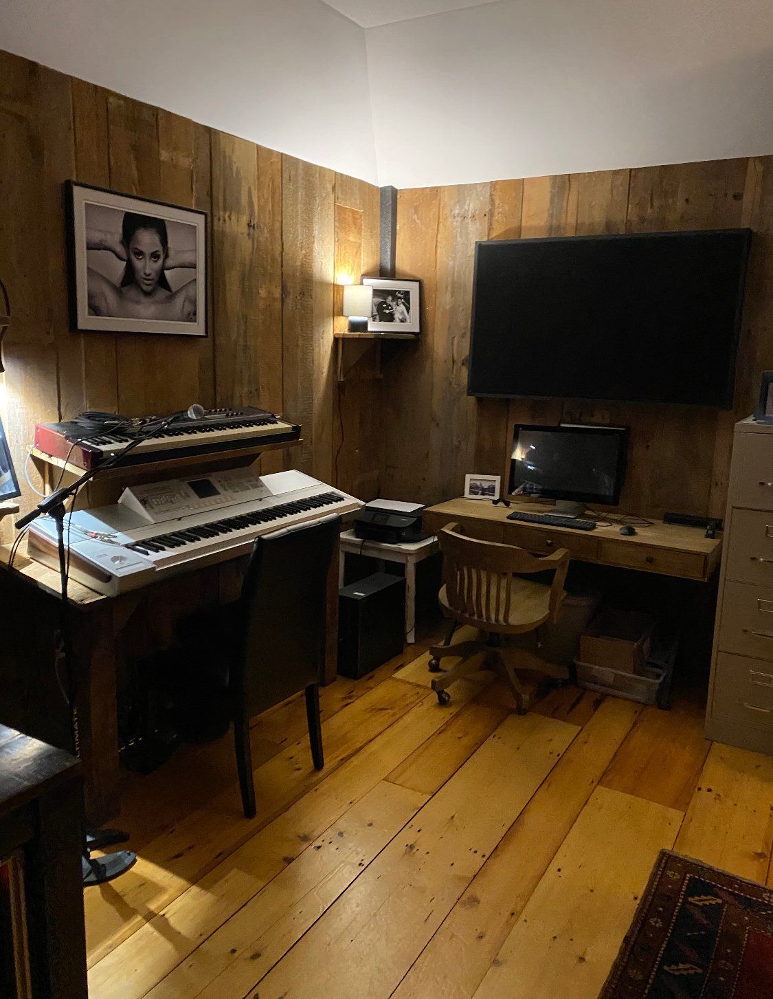Home office with wood-paneled walls and floor; a desk with a keyboard, chair, and computer setup; and a mounted TV.