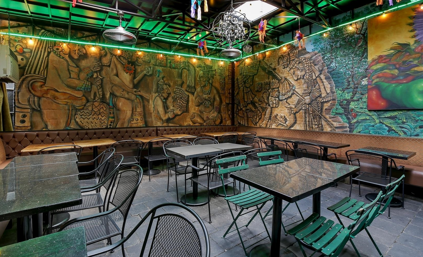 An outdoor dining area with dark tables and green chairs, large mural on the back wall.