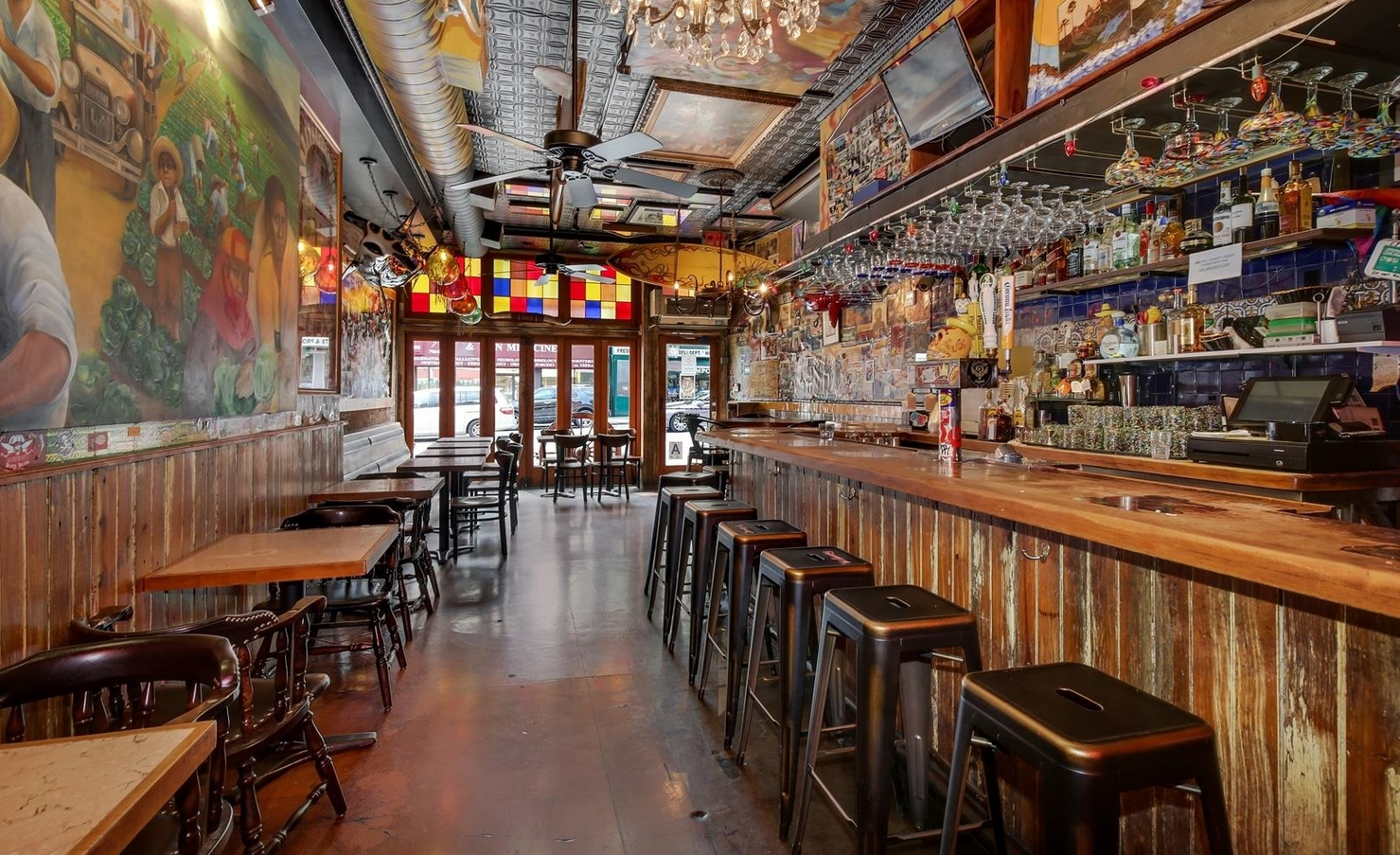 Interior of a dimly lit bar with a long wooden counter, stools, and tables. Mural on the wall, glasses hanging.