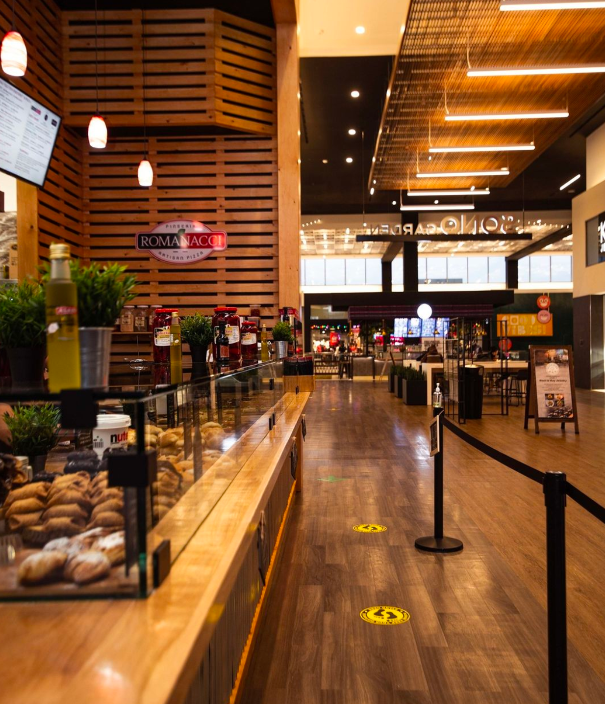 Cafe counter with pastries, plants, and menu, leading to a mall corridor with social distancing markers.