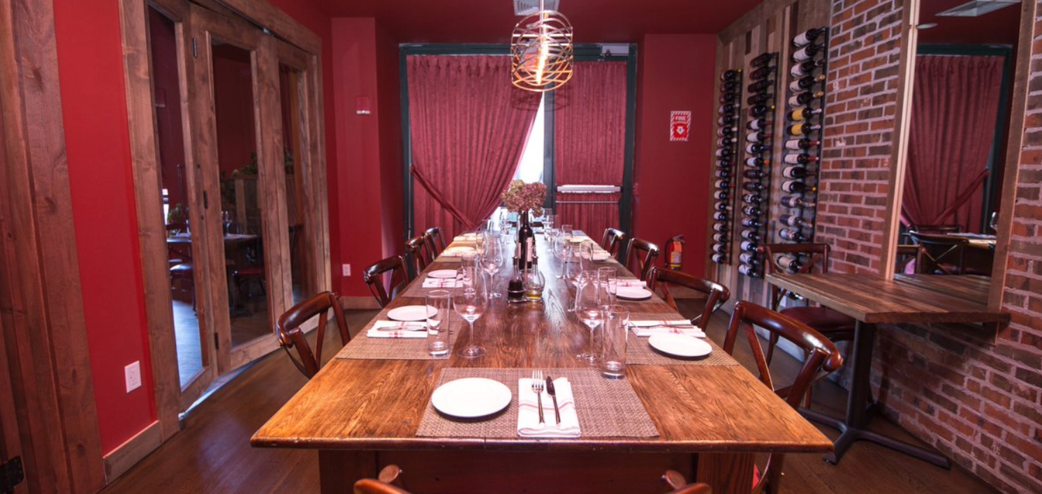A long wooden dining table set in a red-walled restaurant, with place settings and chairs.