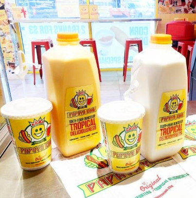 Papaya King drinks and cups on a counter: large jugs and cups of yellow and white beverages with logo.