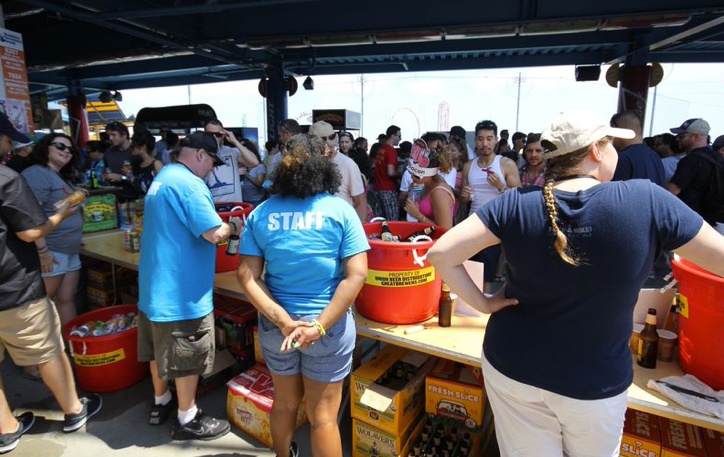 Crowd at an outdoor event with a staff member at a bar serving drinks from red tubs.