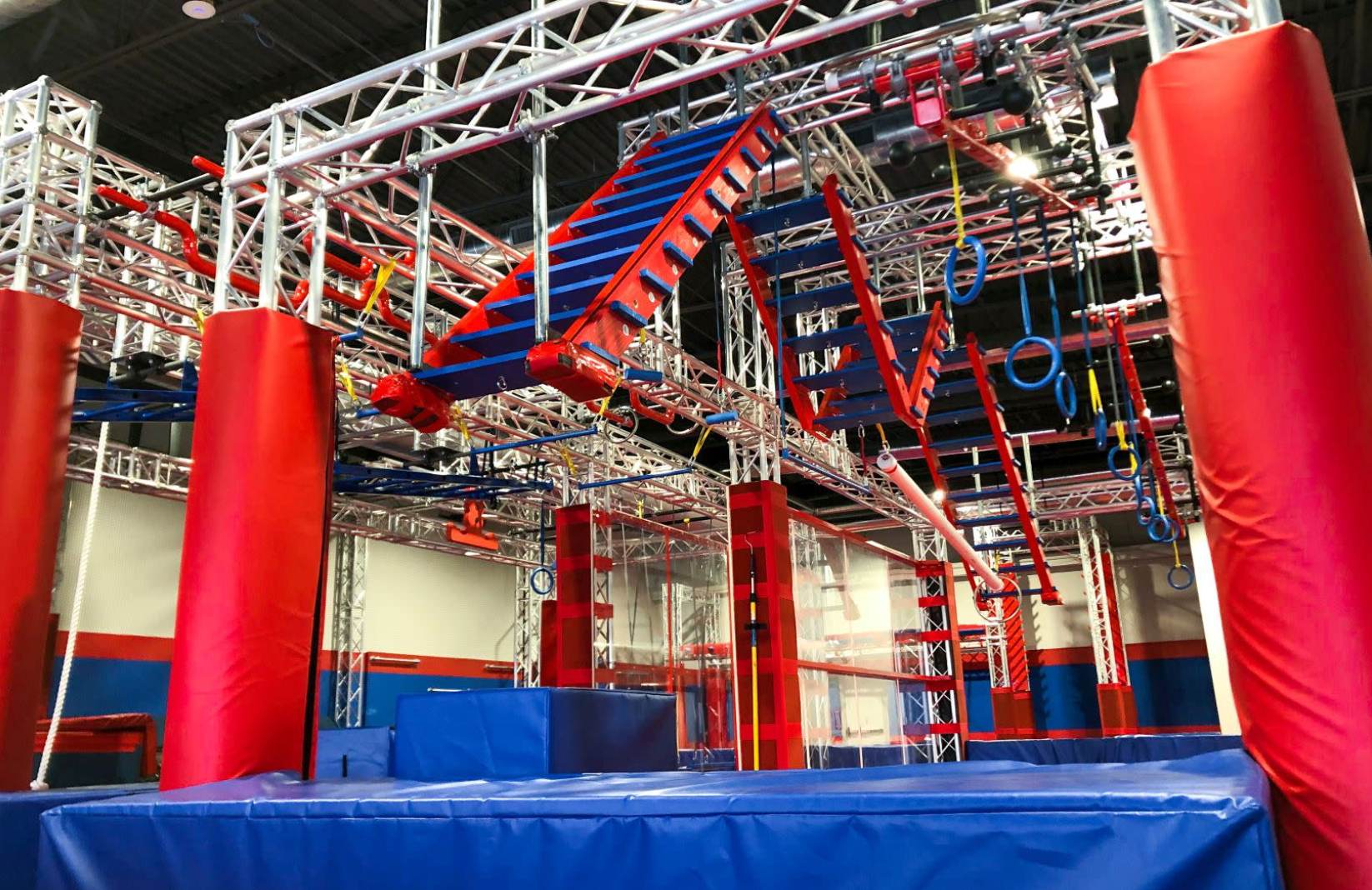 Indoor obstacle course with red, blue, and white structures; includes climbing walls, hanging rings, and padded columns.