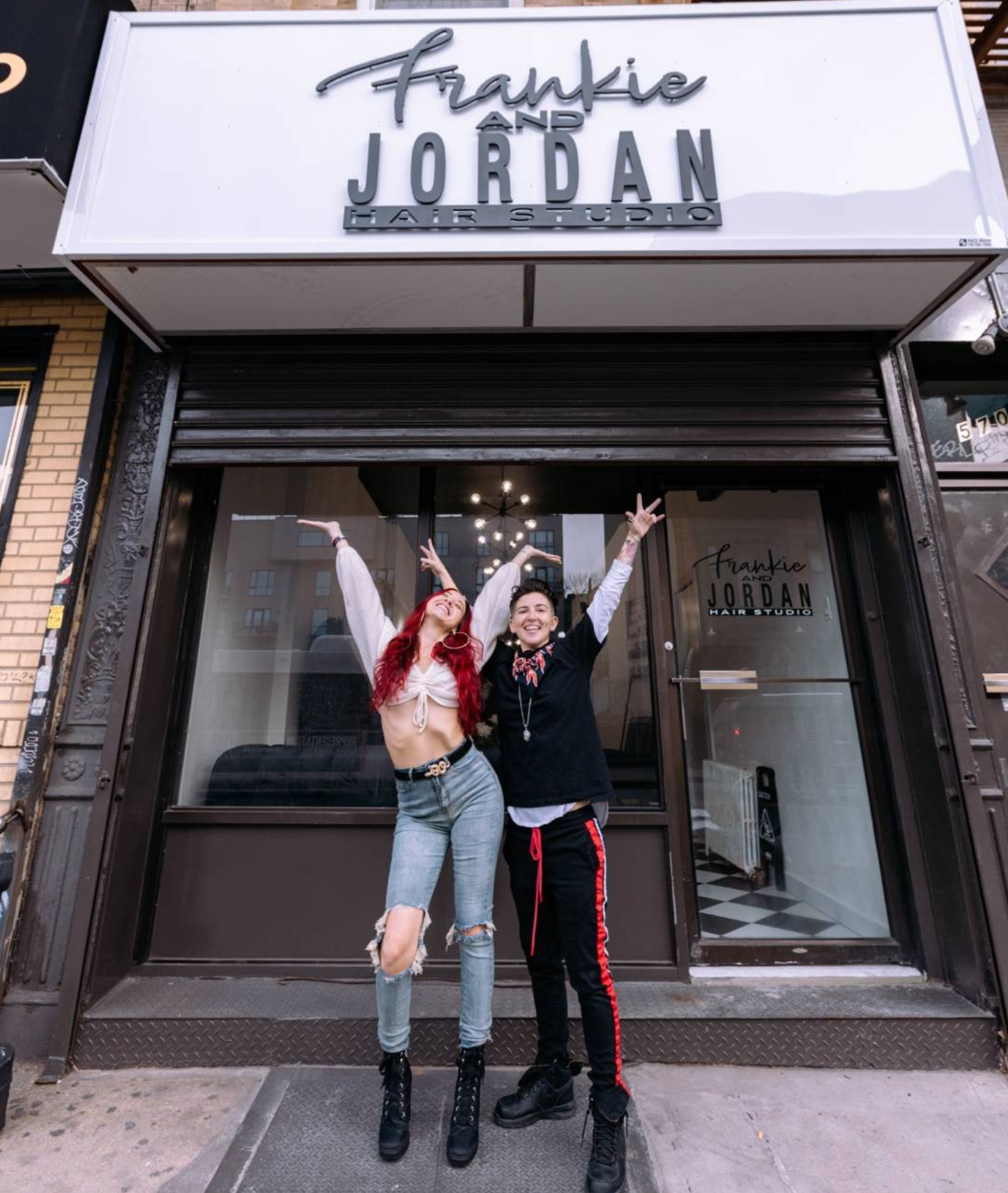 Two people stand in front of a storefront, arms raised. The sign reads 