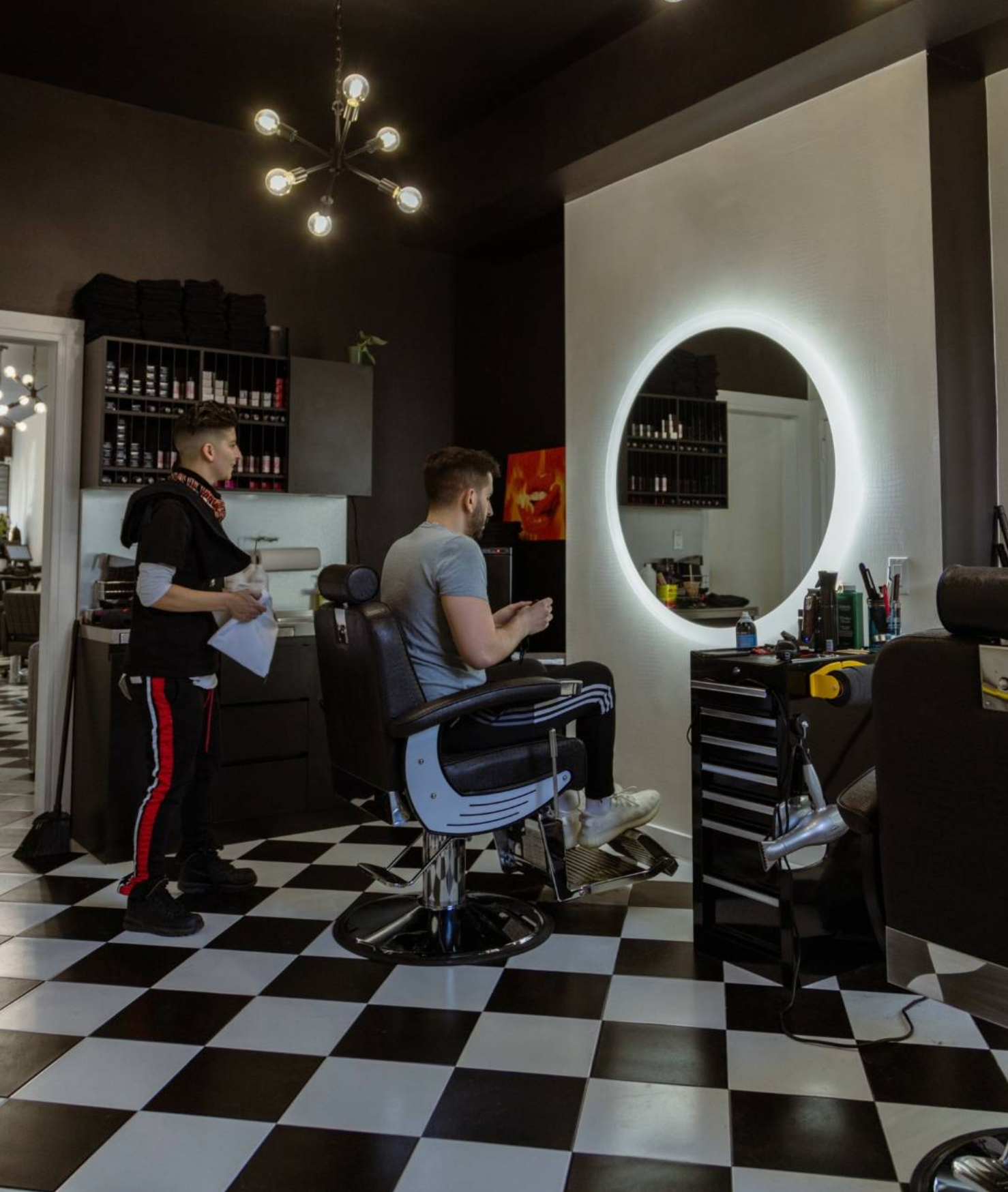 Barbershop interior with client in chair, barber standing, circular mirror with light, checkered floor.