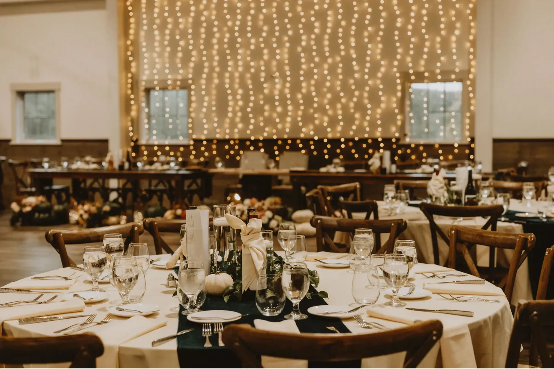 Wedding reception setup with round tables, decorated with centerpieces and linens. Soft lighting and string lights in the background.