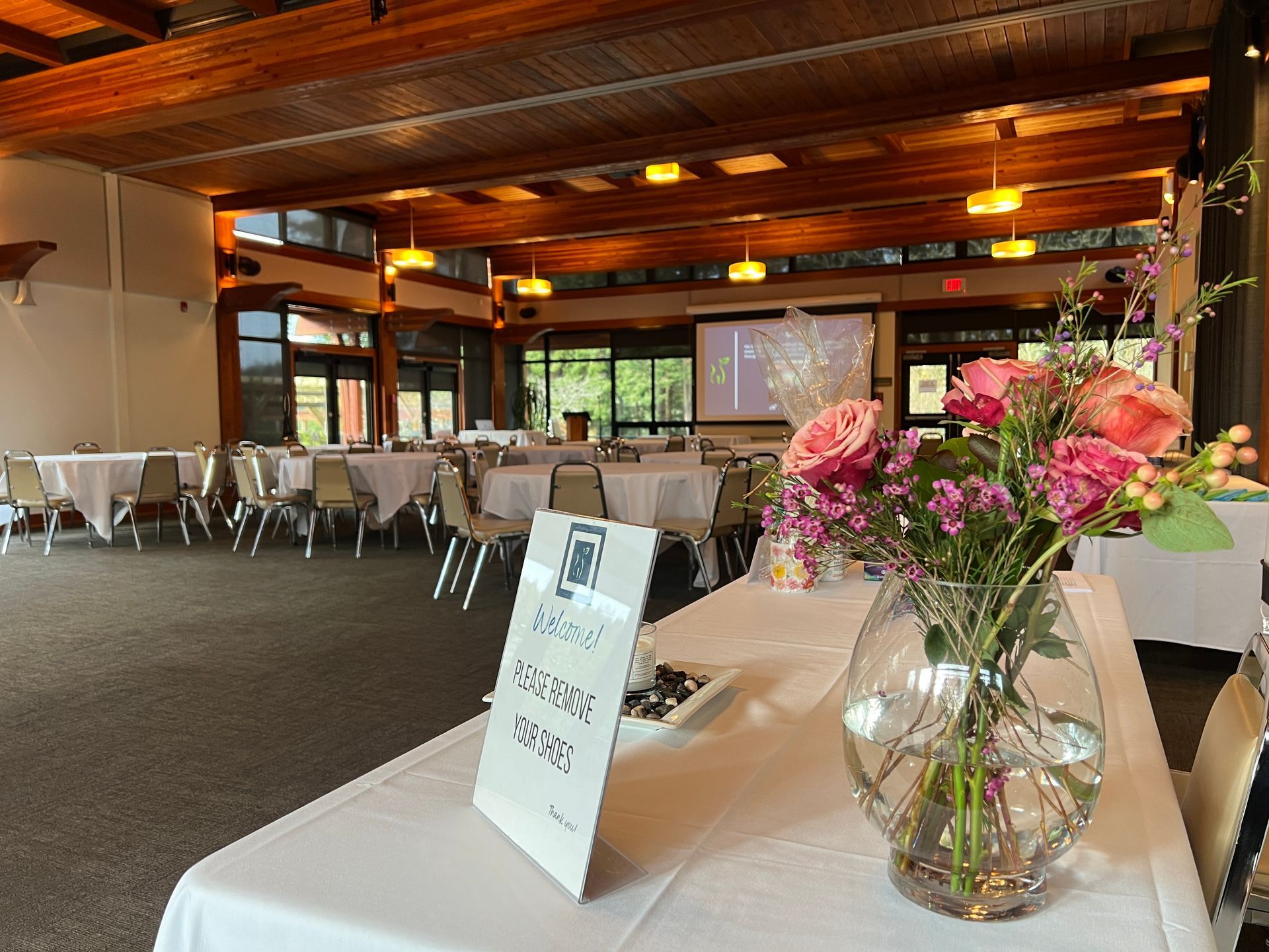 A room set up for an event, with tables covered in white tablecloths. A vase of pink roses is in the foreground.