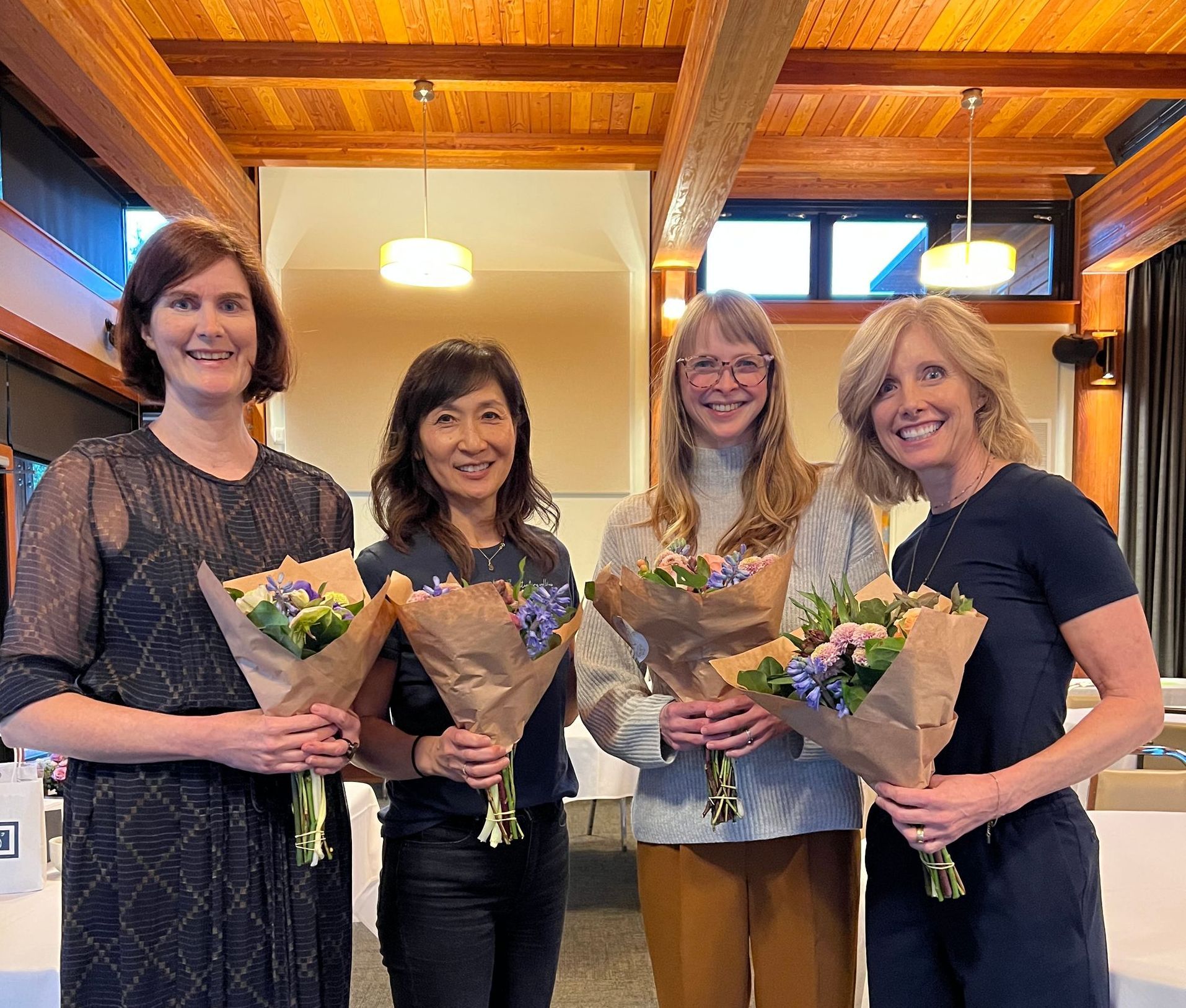 Four women holding bouquets, smiling at the camera, indoors with wood beams, a light-colored wall, and tables.