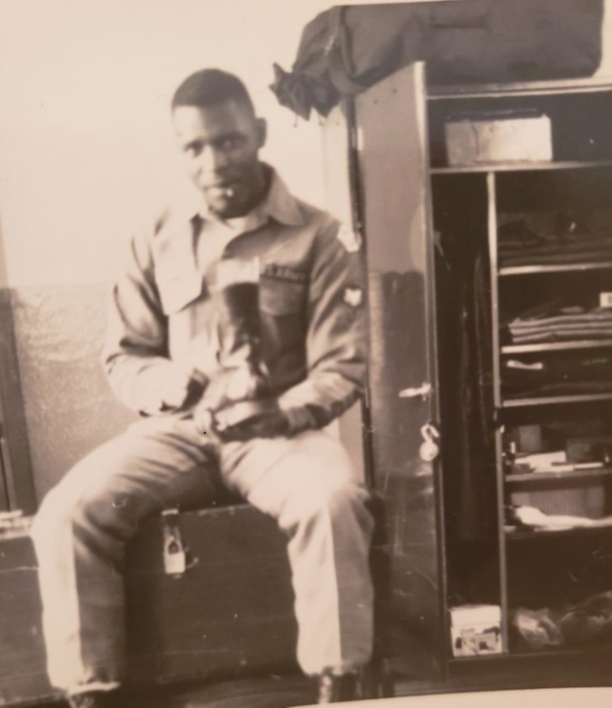 A soldier in uniform sitting on a footlocker next to an open locker, holding a boot while looking toward the camera.