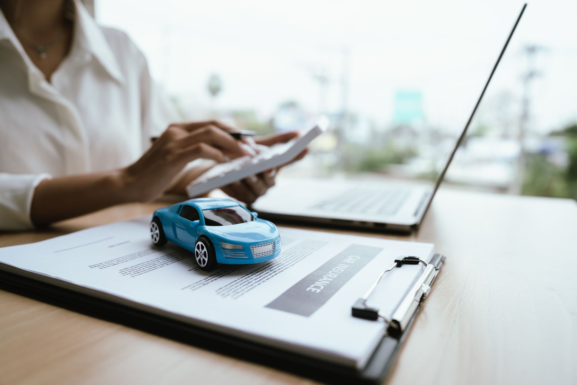 Woman calculating car insurance costs with toy car and policy documents on desk.