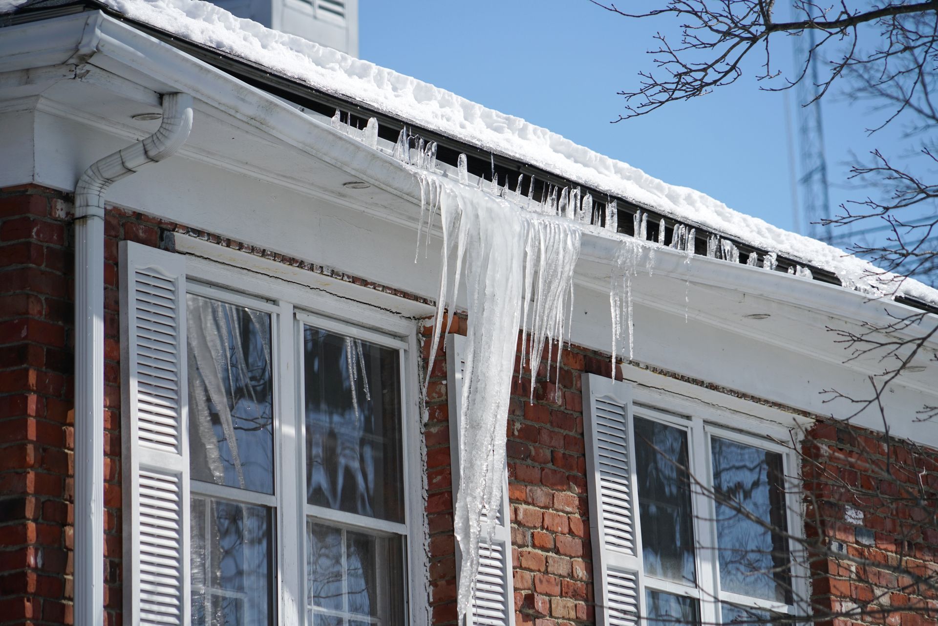Icicles hanging from a snow-covered roof of a brick building with white shutters.