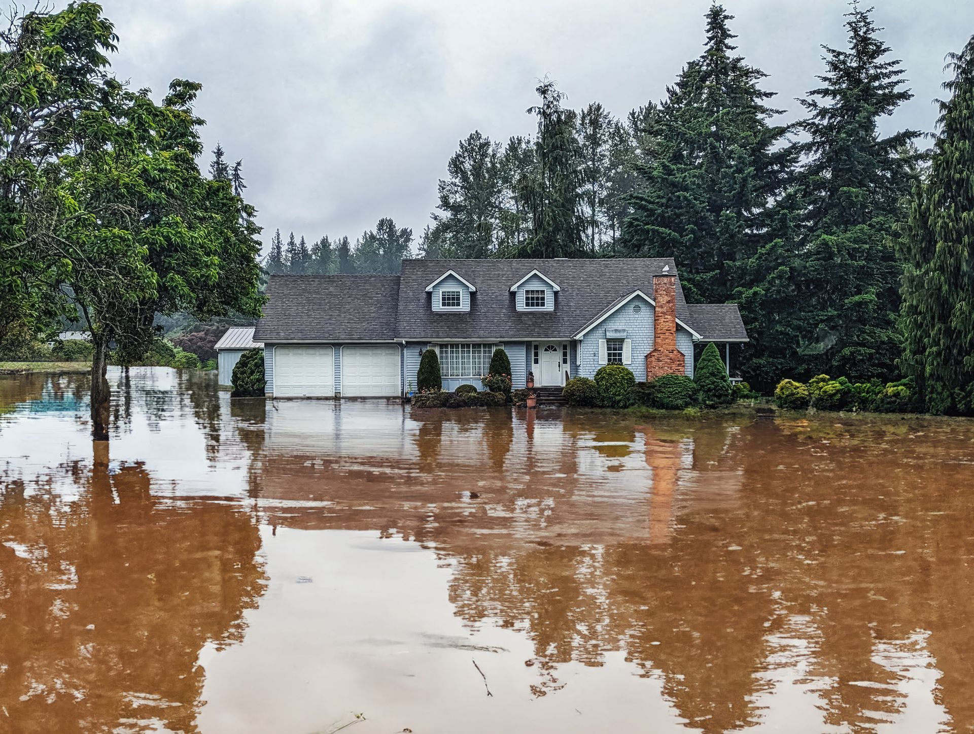 House flooded with brown water, surrounded by trees, on an overcast day.