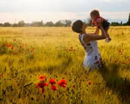 Woman holding up child in a golden field with red flowers, sunlit.