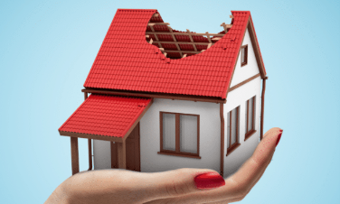 Hand holding a miniature house with a damaged roof, red tiles and white walls, against a blue background.