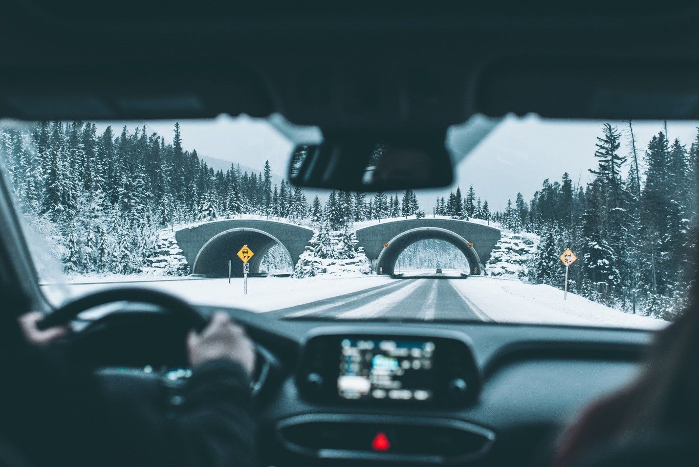 Driver's view through windshield of a car, snowy road, approaching two arch bridges, flanked by snow-covered trees.