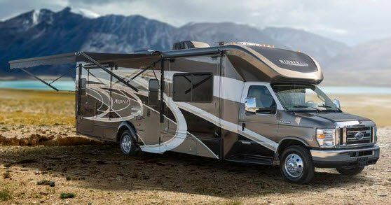 Brown and white RV parked outdoors near a lake and mountains, awning extended.