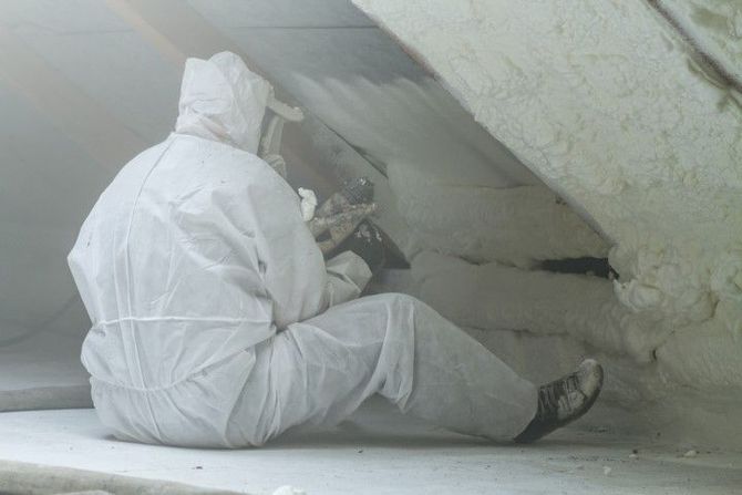 Person in white protective suit inspecting insulation inside a dusty crawlspace