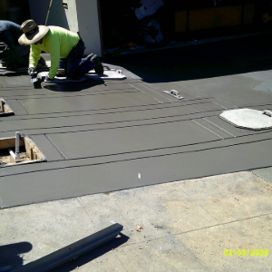 Worker kneeling on a gray rooftop, installing materials near an opening in bright sunlight.