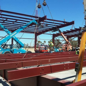 Workers assembling a steel building frame with cranes and a blue lift under a clear sky