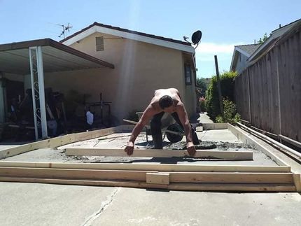 Person bending over lumber in a sunlit driveway beside a house and fence